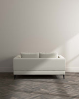 Minimalist modern light cream sofa with clean lines and elegant black tapered wooden legs set against a neutral-toned wall on a herringbone-patterned wooden floor.