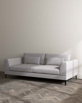 Modern minimalist light grey sectional sofa with clean lines, featuring a chaise lounge on the right side, textured fabric upholstery, straight armrests on either end, low-profile cushioned backrest, and elegant tapered black legs. Set against a neutral wall on a herringbone-patterned wood floor.