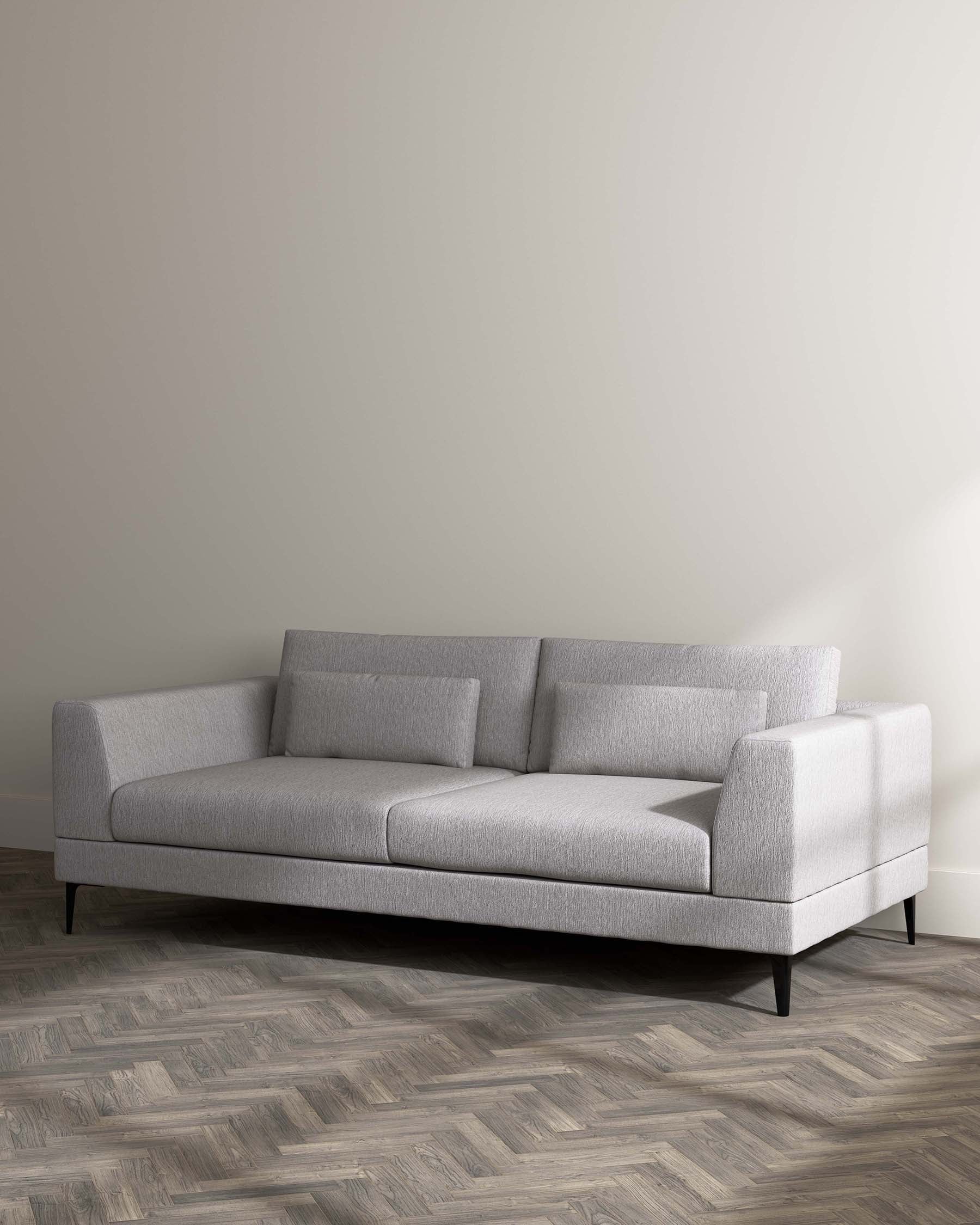 Modern minimalist light grey sectional sofa with clean lines, featuring a chaise lounge on the right side, textured fabric upholstery, straight armrests on either end, low-profile cushioned backrest, and elegant tapered black legs. Set against a neutral wall on a herringbone-patterned wood floor.