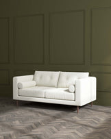 Modern minimalist three-seater sofa with a clean design, featuring plush ivory upholstery and cylindrical side cushions. The sofa has a low backrest, simple lines, and stands on splayed round wooden legs in a darker hue that contrasts with the light fabric.