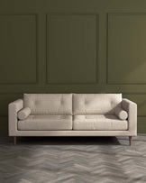 Modern beige fabric sofa with clean lines and cylindrical bolster pillows, set against a dark green wall with panel detailing, standing on a herringbone patterned wooden floor.