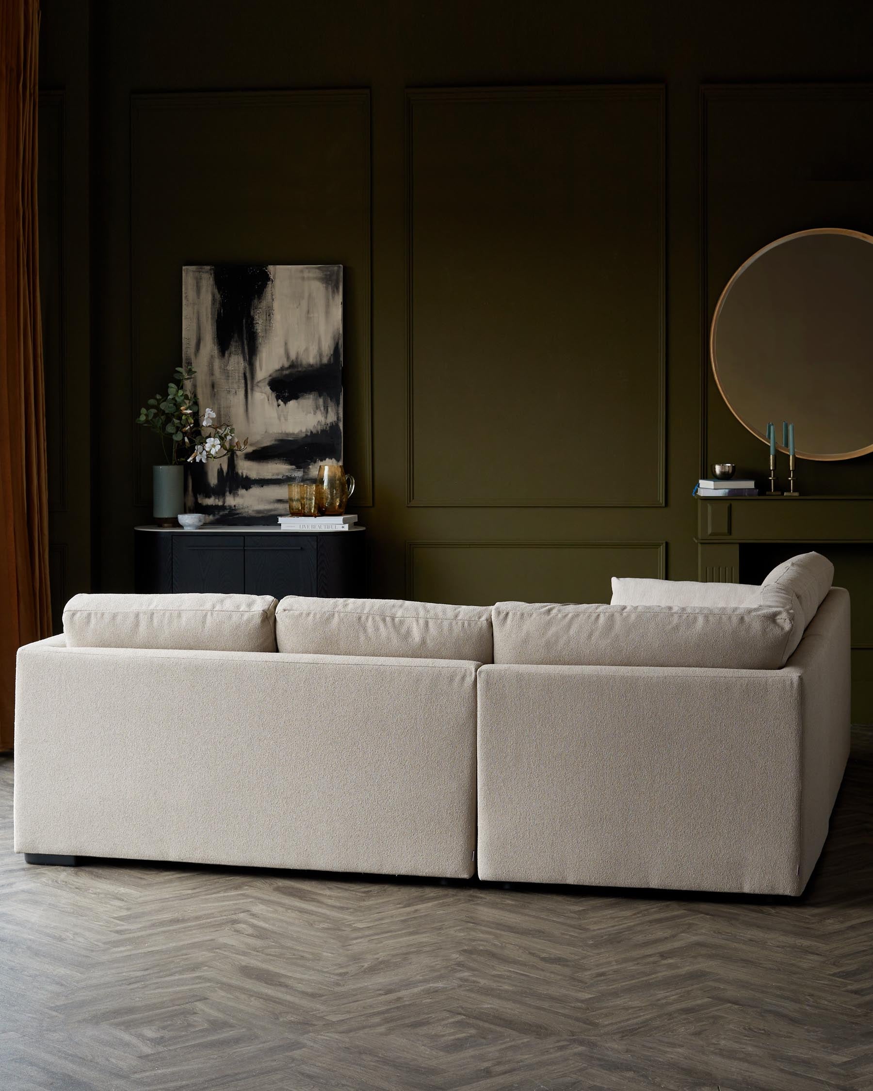 A contemporary beige modular sofa with clean lines and plush cushions, on a dark herringbone floor against a backdrop of green panelled walls with decorative items, including a painting, round mirror, and sideboard.