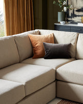 Contemporary L-shaped sectional sofa in a neutral beige fabric with plush back cushions and three accent pillows in earth tones. A sleek black sideboard table with a vase and decor pieces is in the background.