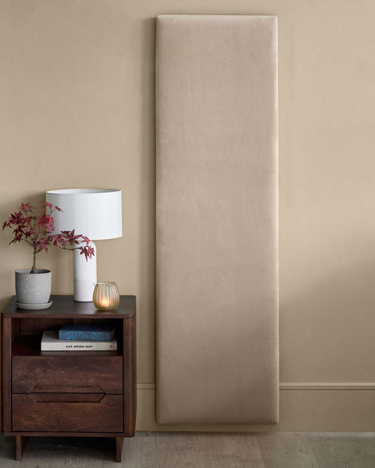 A modern nightstand with dark wood finish, lamp, decorative plant, and plush beige wall panel against a neutral backdrop.