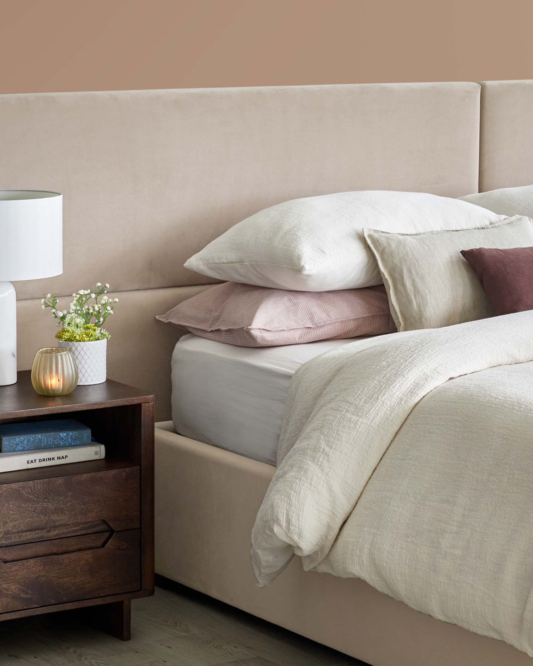 A cozy bed with layered pillows, a textured duvet, and a dark wood nightstand featuring decorative items and a lamp.