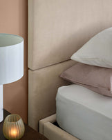 A stylish bed with a soft beige headboard, layered pillows, and a sleek bedside lamp next to a warm candlelight.