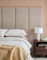 Soft beige upholstered headboard, plush bedding with varied textures, a wooden nightstand, and a modern lamp with plant accents.