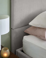 A soft gray upholstered headboard, layered pillows, and a bedside table with a lamp and decorative candle.