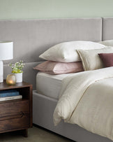 Stylish bed with plush gray headboard, layered bedding, and a modern nightstand featuring a lamp and decorative accents.