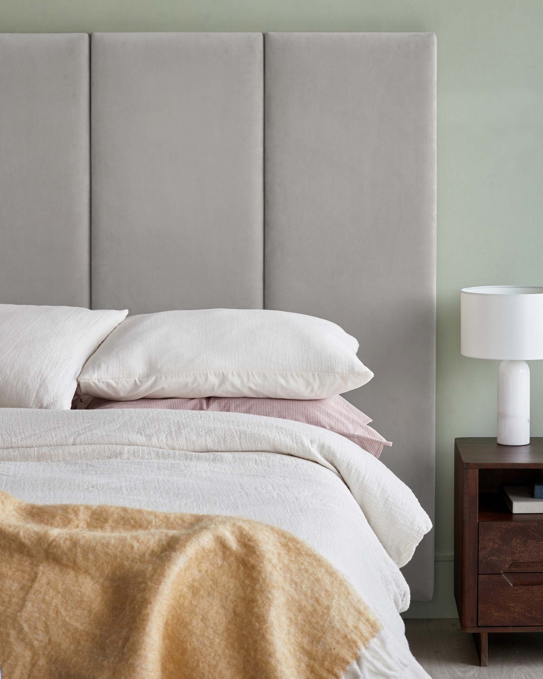 Stylish gray upholstered headboard, layered bedding with white and pastel tones, and a modern nightstand with a sleek lamp.