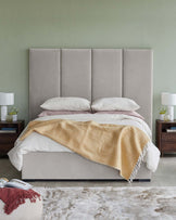 A modern bed with a padded gray headboard, layered bedding, wooden nightstands, and decorative lamps on either side.