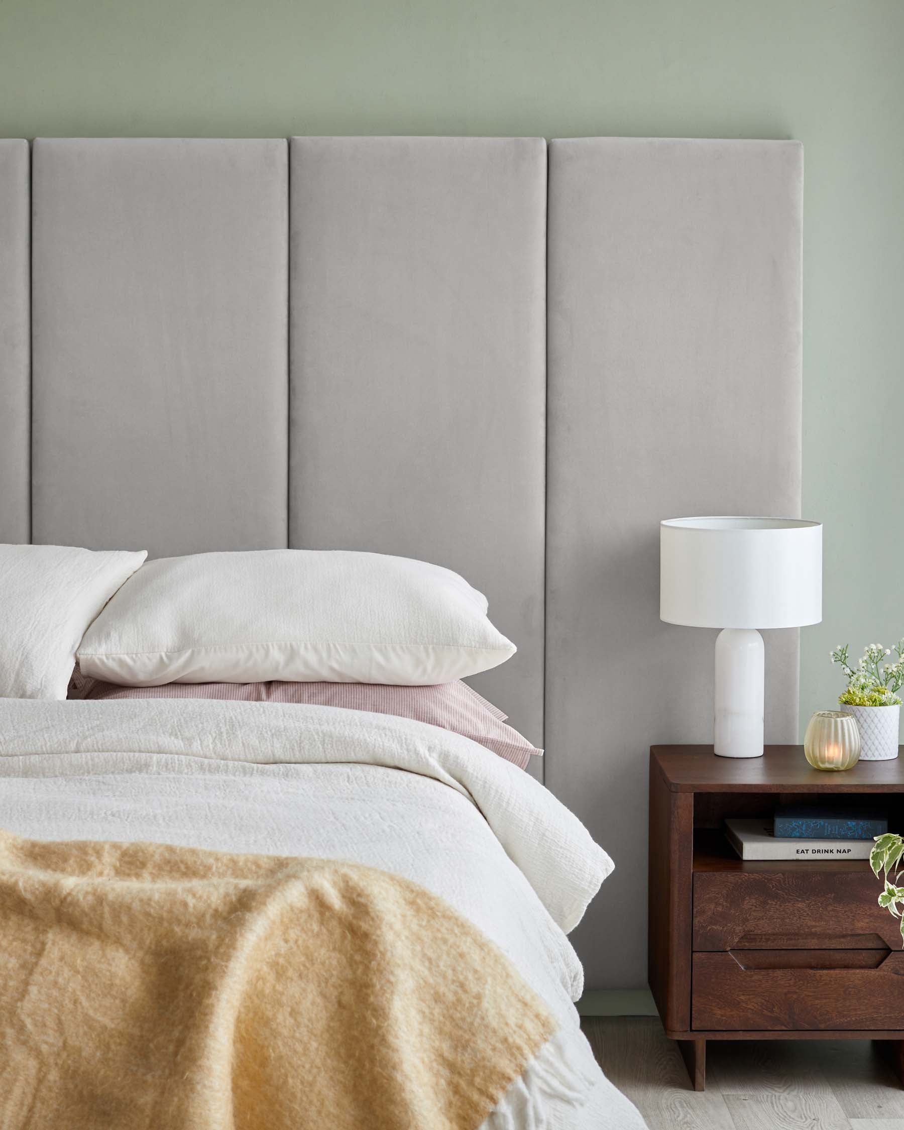 Soft gray upholstered headboard, layered bedding, wooden nightstand with a lamp and plants, creating a cozy bedroom atmosphere.