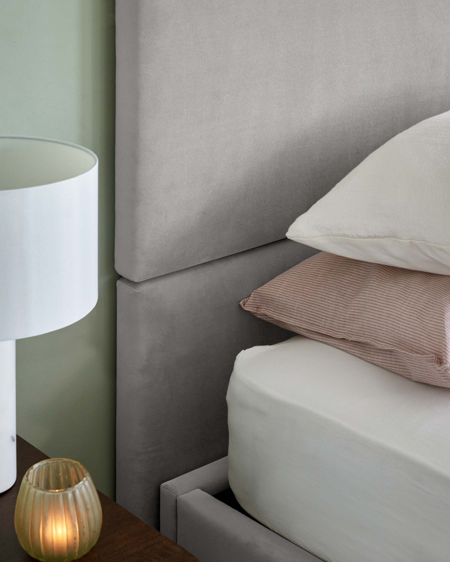 Gray upholstered bed with stacked pillows, bedside table lamp, and a decorative glass candle holder.