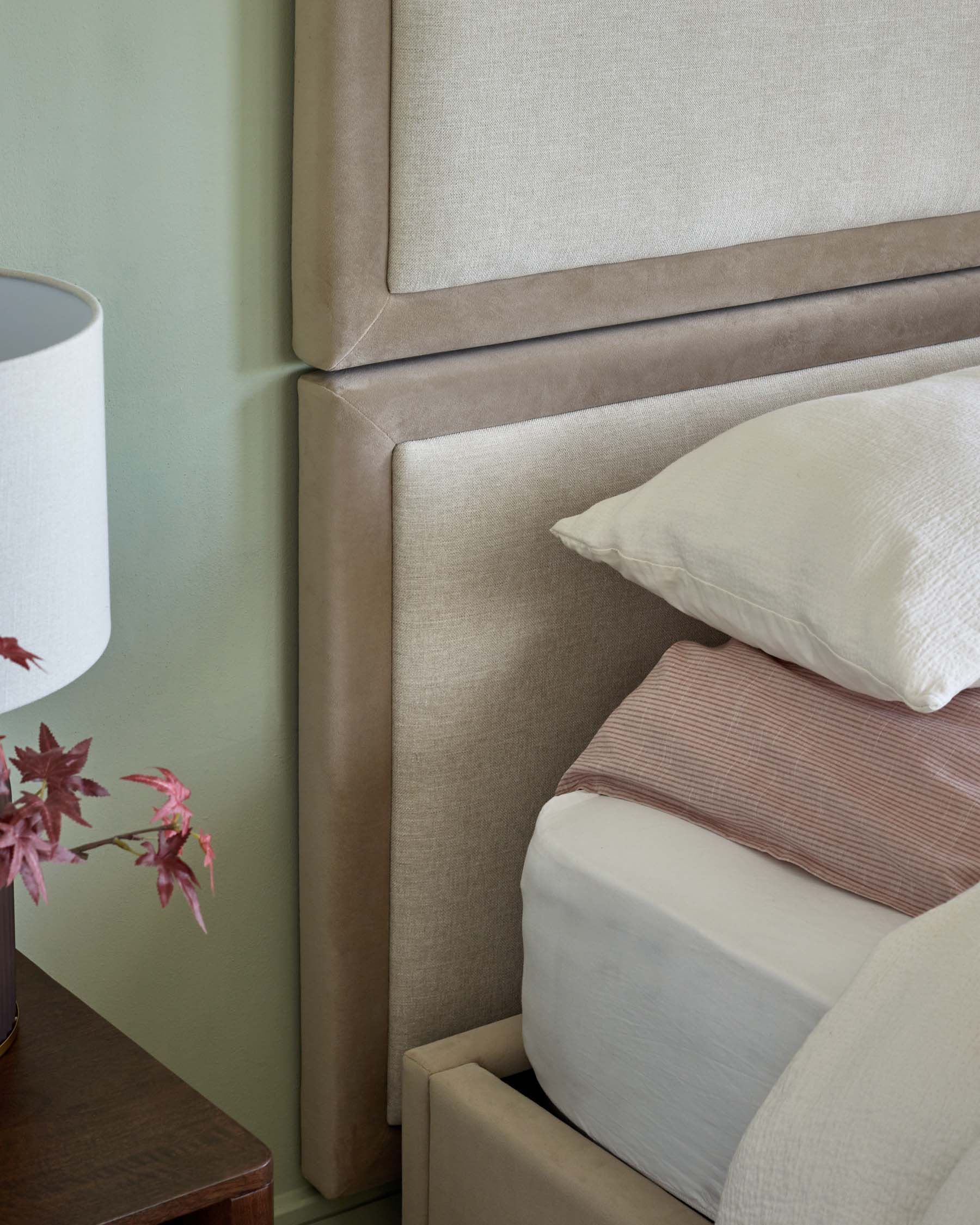 Elegant upholstered bed with a tall, soft headboard, layered bedding, and a bedside table featuring a lamp and decorative plant.