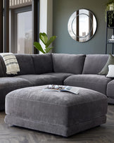 Stylish grey sectional sofa with a matching plush ottoman, accented by a decorative pillow and a modern circular mirror on the wall.