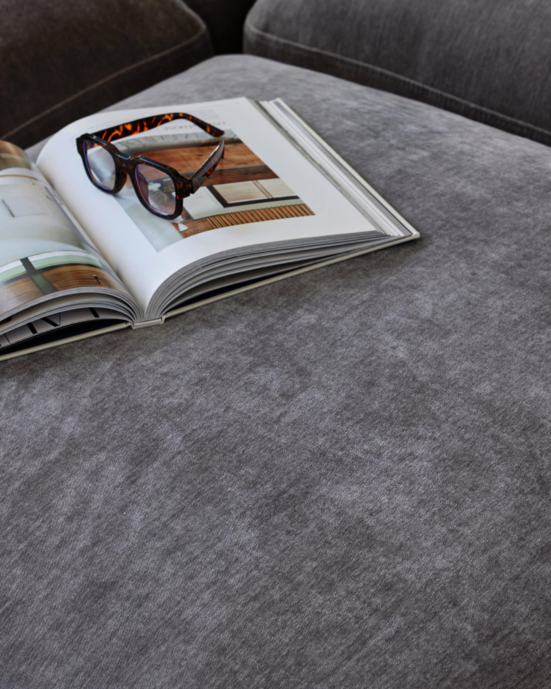 A soft grey upholstered surface featuring an open magazine and stylish glasses, creating a chic, inviting atmosphere.