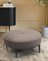 A round, padded ottoman in beige with a button detail, paired with a sleek black cabinet and a decorative plant in a pot.