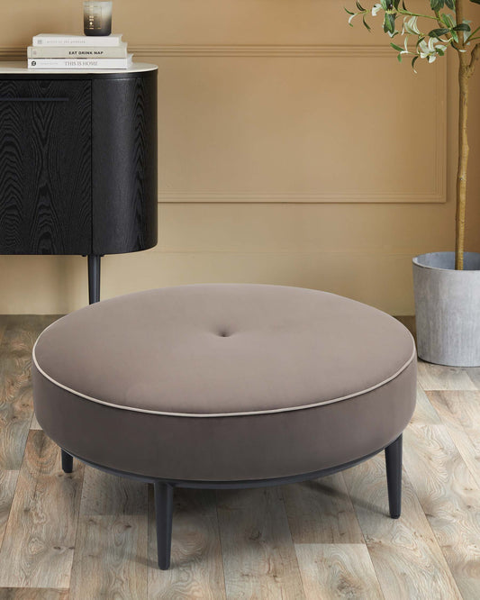 A round, padded ottoman in beige with a button detail, paired with a sleek black cabinet and a decorative plant in a pot.