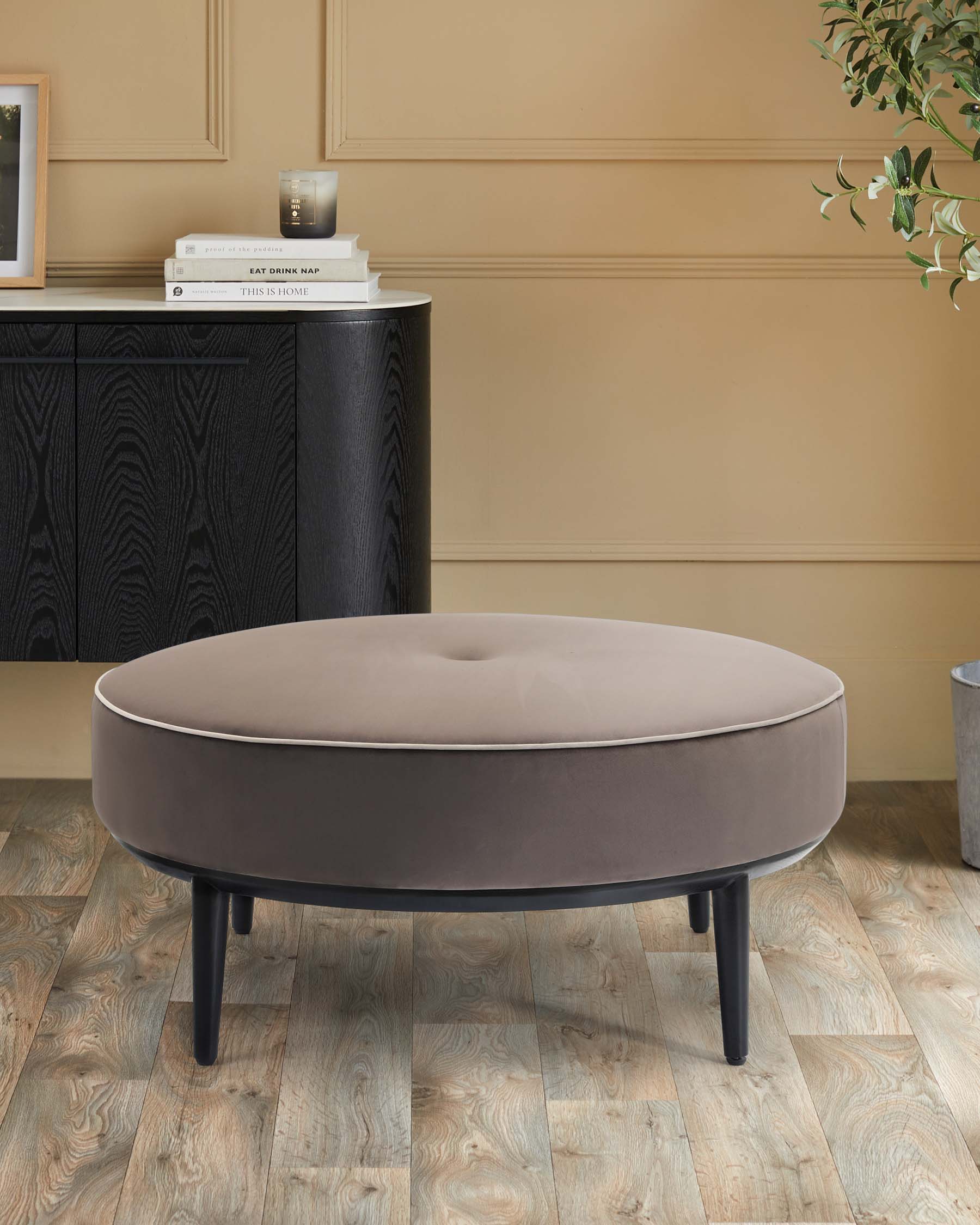 Round, tufted ottoman in soft grey fabric with a slim black base, paired with a sleek dark cabinet and decorative accessories.