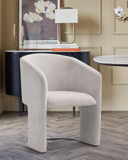 A sleek, modern chair with a curved design in soft beige fabric, placed by a stylish lamp and decorative table.