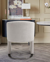 A soft, cream upholstered chair with sleek metal legs, a modern lamp, and stylish decor on a round table against a neutral wall.