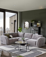 A modern gray sectional sofa, circular glass coffee tables, a decorative plant, and minimalist shelving against a green wall.