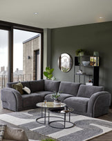 Stylish gray sectional sofa, round glass coffee tables, and a black shelving unit, accented by greenery and a circular mirror.