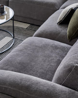 A plush gray sectional sofa with soft cushions, paired with a sleek round glass coffee table and a decorative pillow.