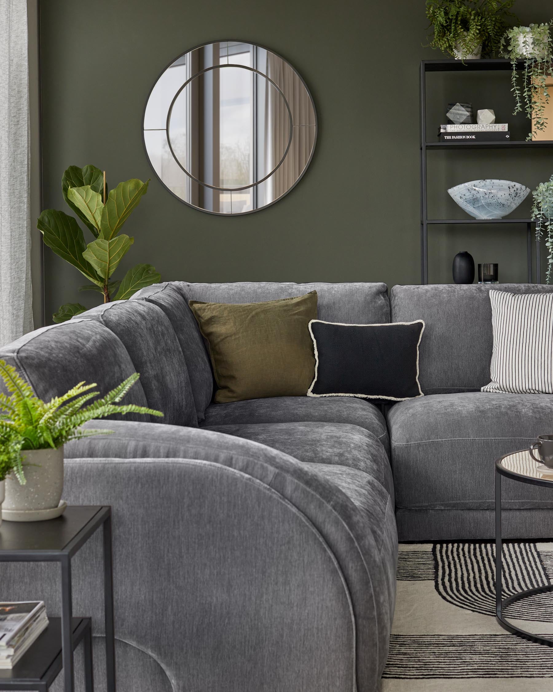 Gray sectional sofa with decorative pillows, round mirror, black metal shelving, potted plant, and a small side table.