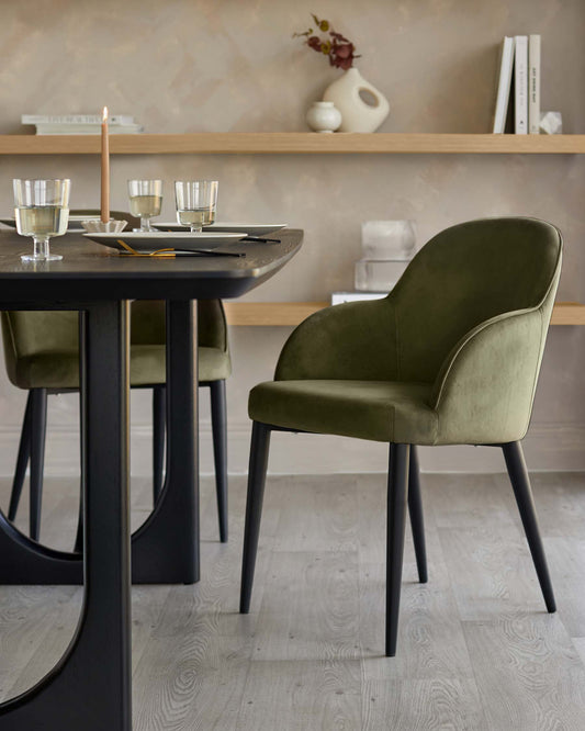 Sleek black dining table paired with stylish green velvet chairs, set with elegant plates and glasses, against a subtle backdrop.