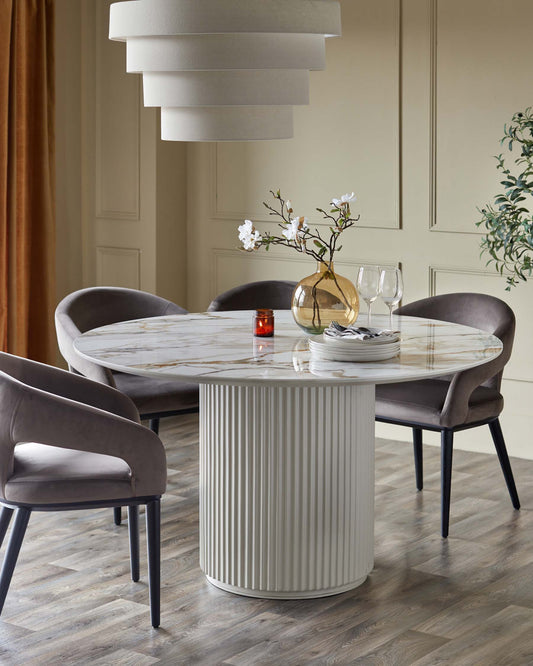 A round marble dining table with a fluted base, paired with plush gray chairs, accentuated by decorative vases and tableware.