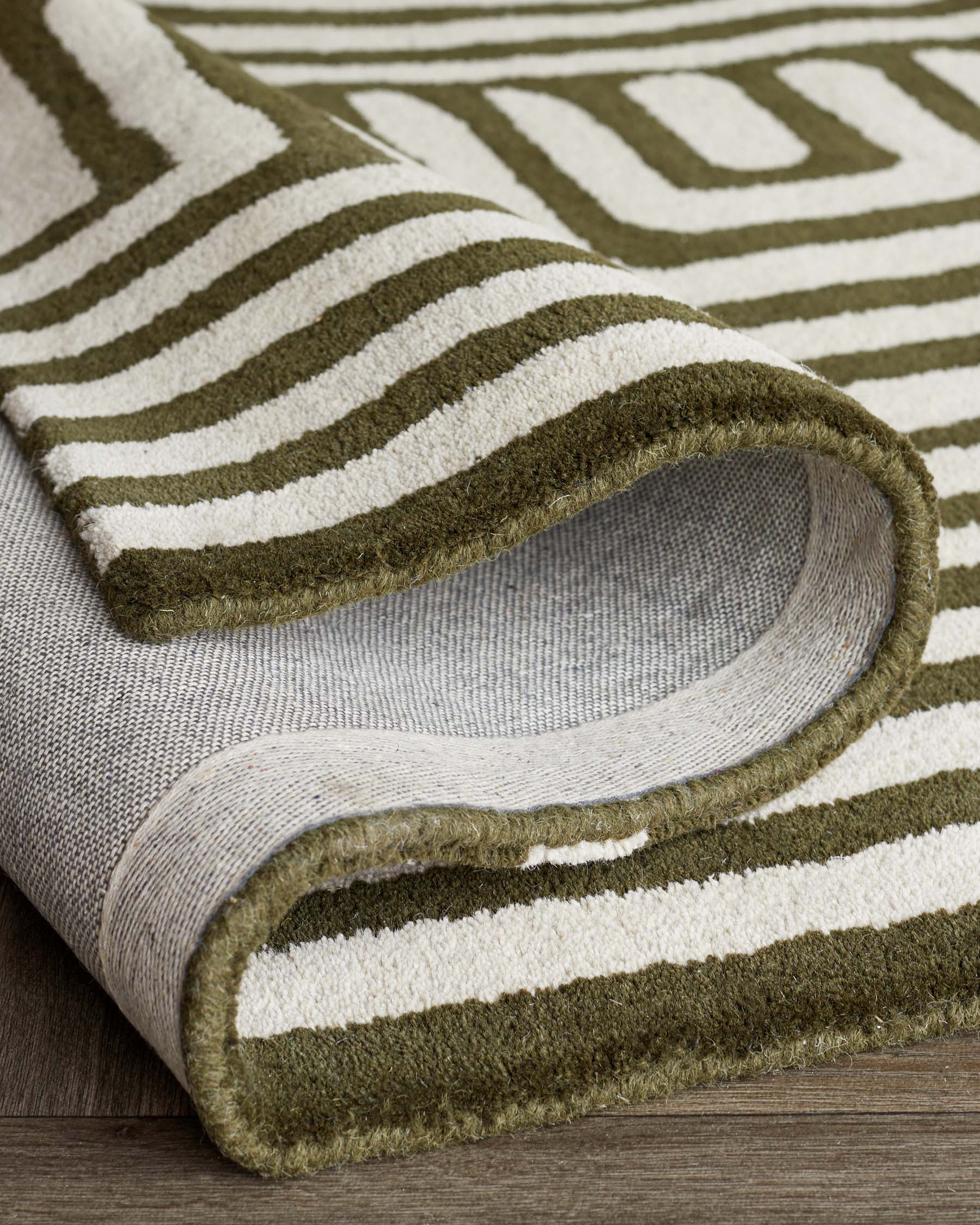 Close-up of a contemporary striped area rug with olive green and cream colours, showcasing its texture and folded corner to reveal the underside, positioned on a wooden floor.