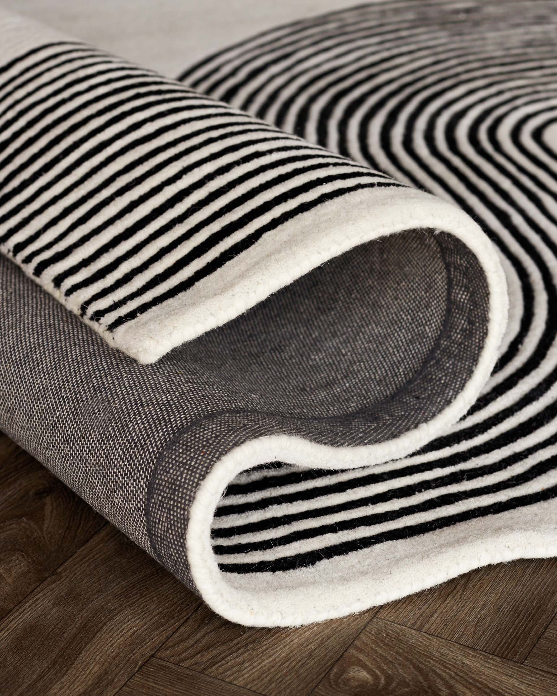 A modern area rug with a black-and-white striped pattern, rolled up at the corner to reveal its texture, against a wooden floor background.