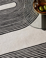 Round glass-top side table with a metallic base, accompanied by a wooden tray with metallic details holding a small silver vase and fruit. The table rests on a textured rug with linear patterns.