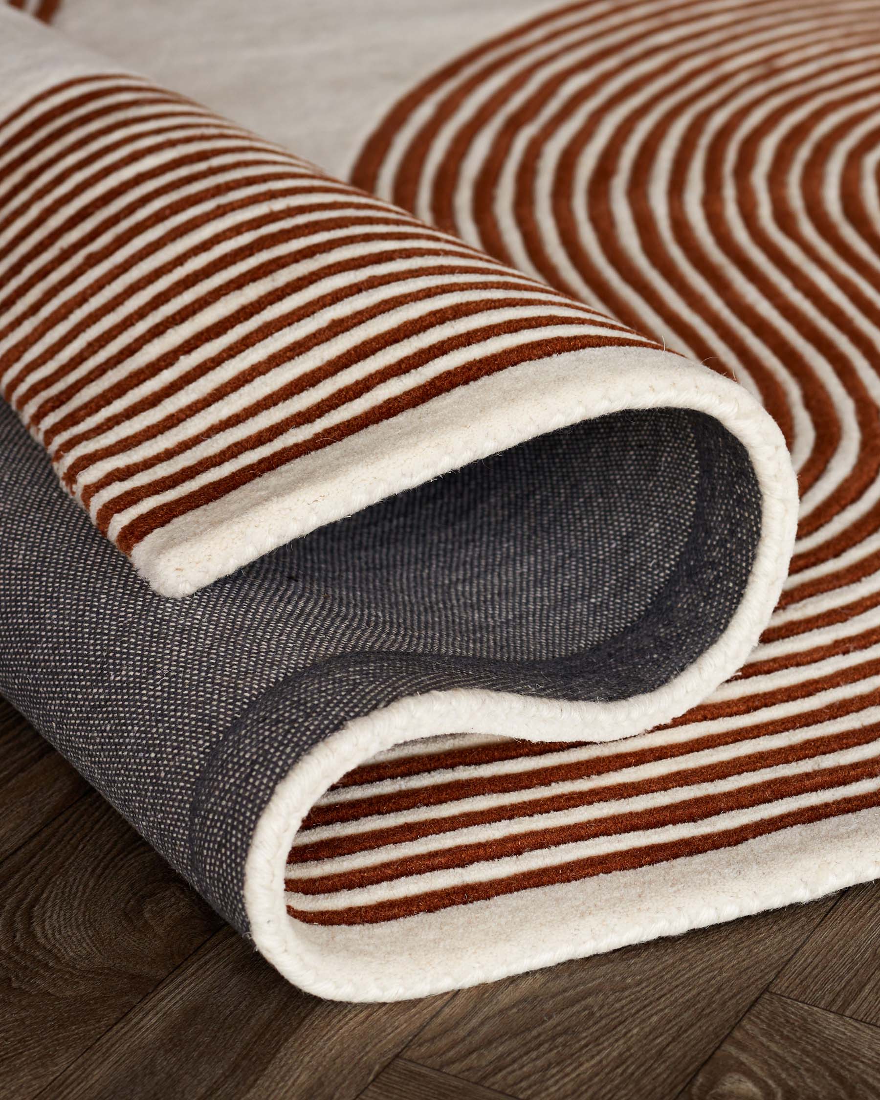 A rolled-up area rug with a brown and white striped pattern on top of a textured dark grey rug on a wooden floor.