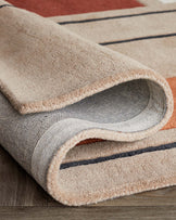 Contemporary area rug with a rolled-up corner revealing a textured underside, featuring neutral beige tones with bold red and blue stripe accents on a wooden floor.