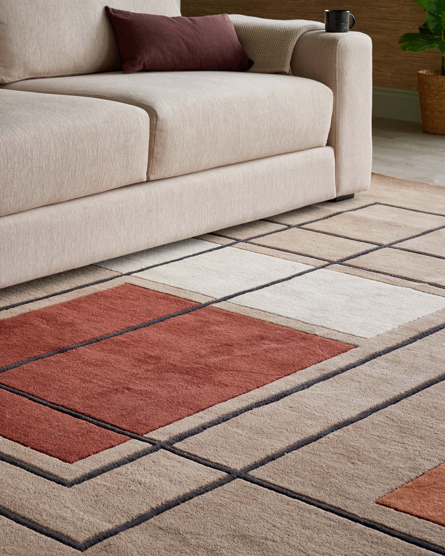 Beige upholstered sofa with clean, straight lines and plush cushioning, complemented by a textured throw pillow in earthy tones, set atop a contemporary area rug featuring a geometric pattern in shades of rust, beige, and cream.