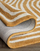 Striped area rug with beige and white pattern, rolled at one end to show texture and thickness, on a wooden floor.