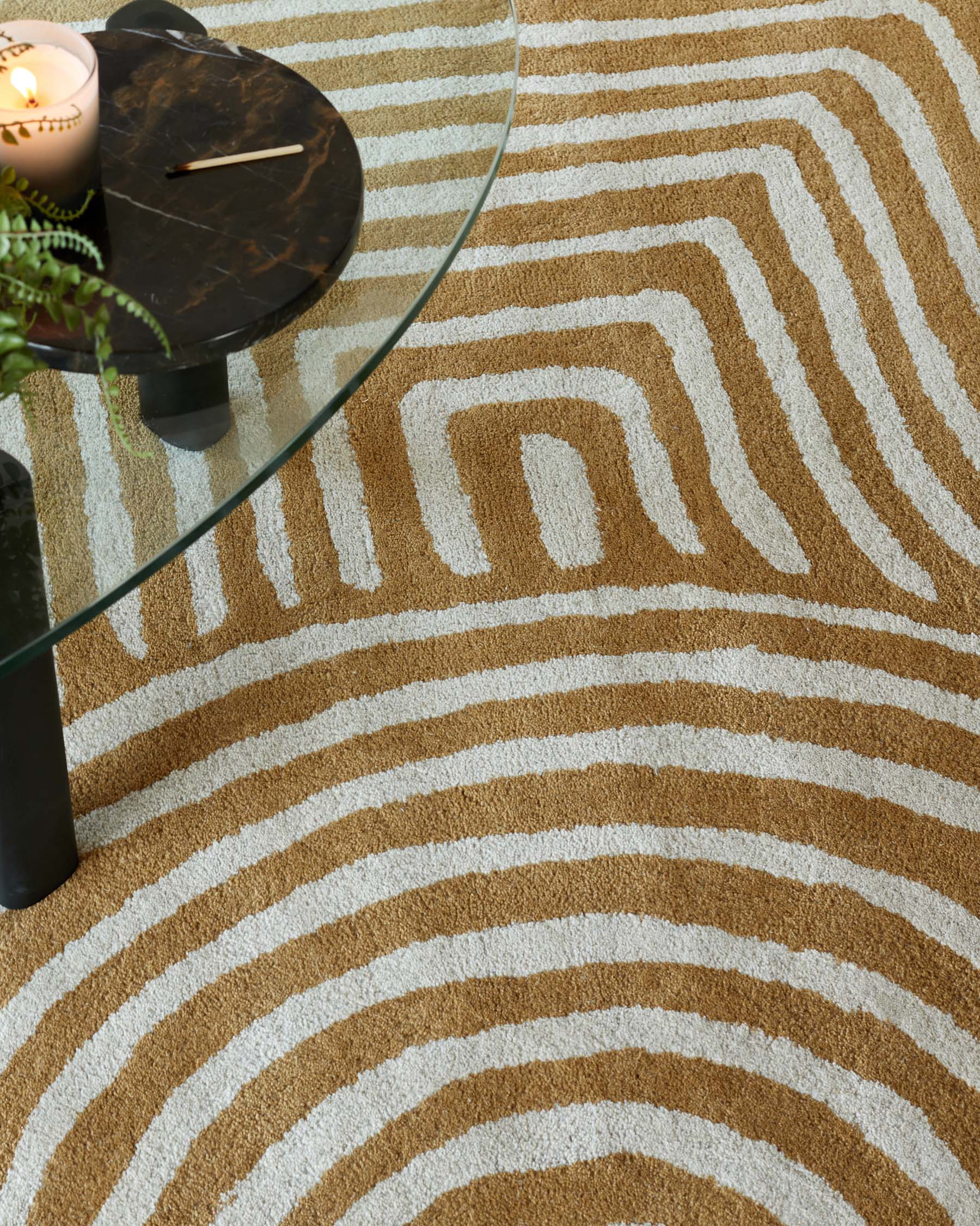 Round glass-top side table with a dark marble base, accompanied by a textured patterned rug featuring earthy tones and concentric geometric shapes.