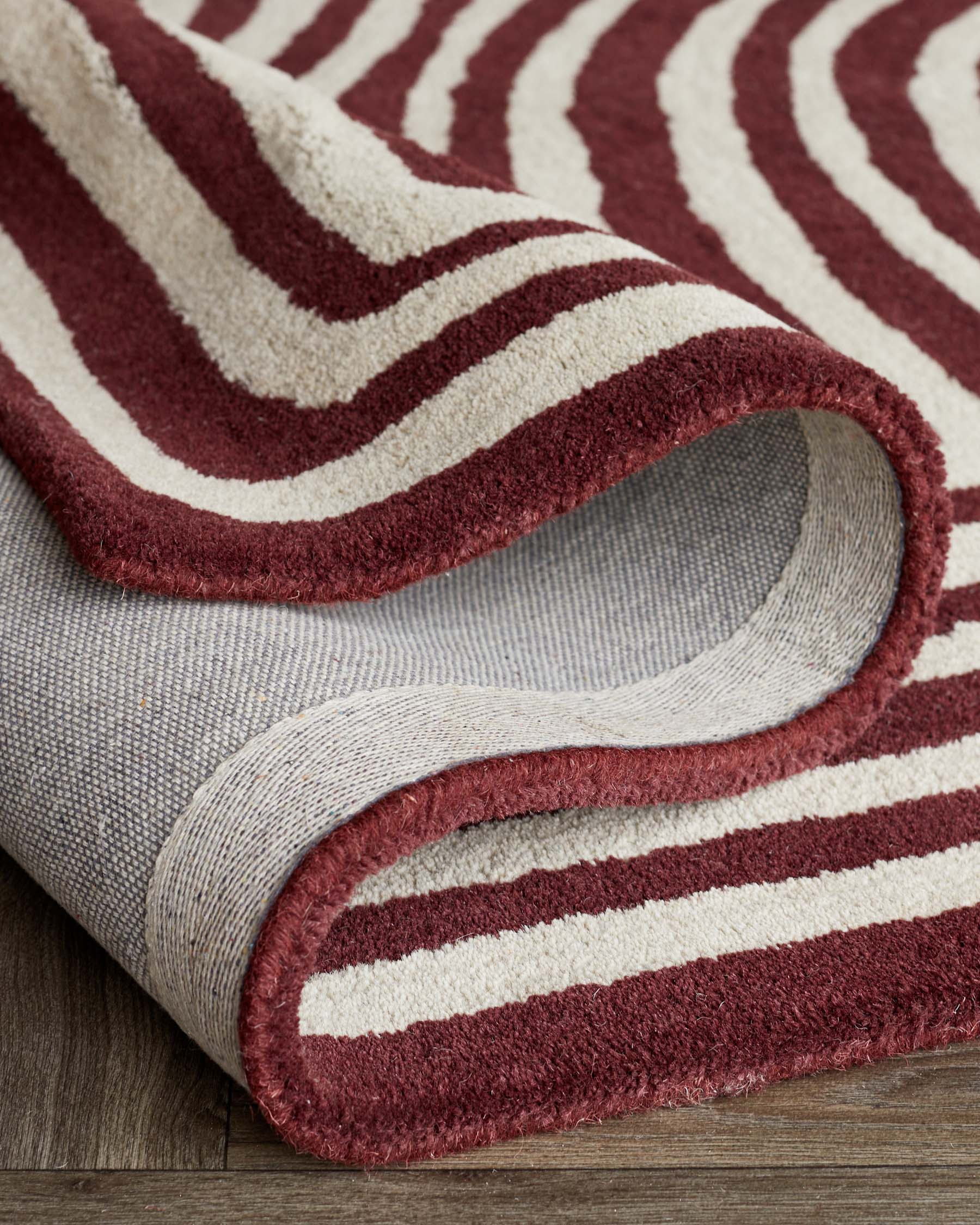 Striped area rug with a curved edge, displaying a pattern of maroon and cream stripes on a textured surface, partially rolled up on a wooden floor.