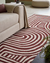 Beige sofa with a cozy throw, a patterned red and cream rug, a small round table, and a soft pouf nearby.