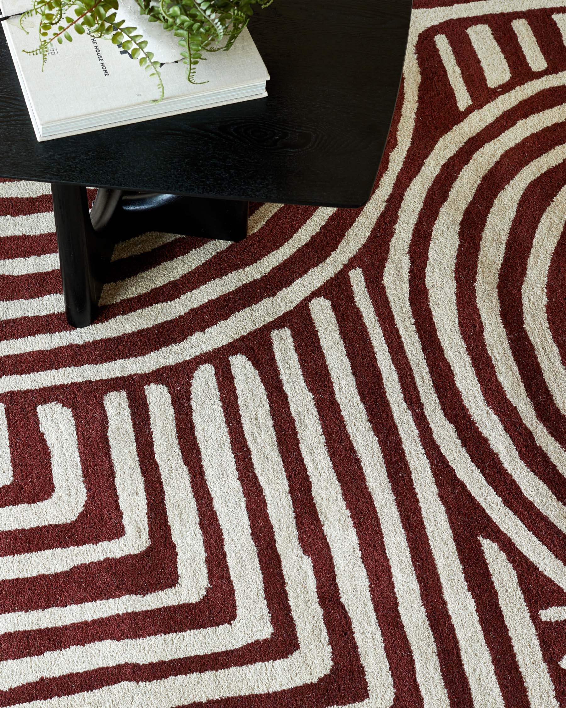 A sleek black coffee table atop a patterned area rug featuring bold maroon and cream waves, complemented by a small green plant.