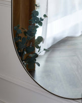 Partial reflection of a sleek, modern oval mirror with a thin metallic frame, mounted on a white wall, featuring a soft reflection of a eucalyptus plant and subtle hints of a light-filled room.