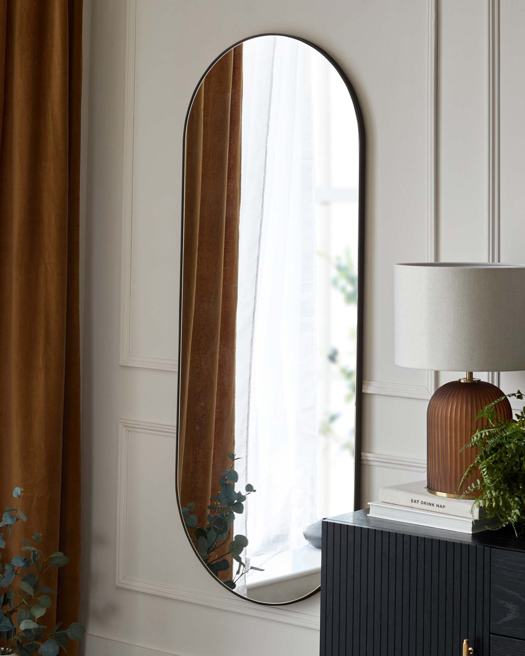 An elegant, tall arched mirror with a slim dark frame leans against a white wall with wainscoting. To the right, a contemporary nightstand or small cabinet with a dark, textured surface and minimalist brass handle serves as a base for a stylish lamp with a fluted, golden base and white shade, accompanied by neatly stacked books and a potted plant.