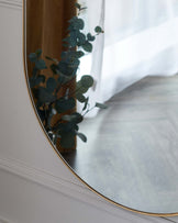 Elegant, wall-mounted oval mirror with a thin golden frame, reflecting a gentle touch of greenery from a houseplant.