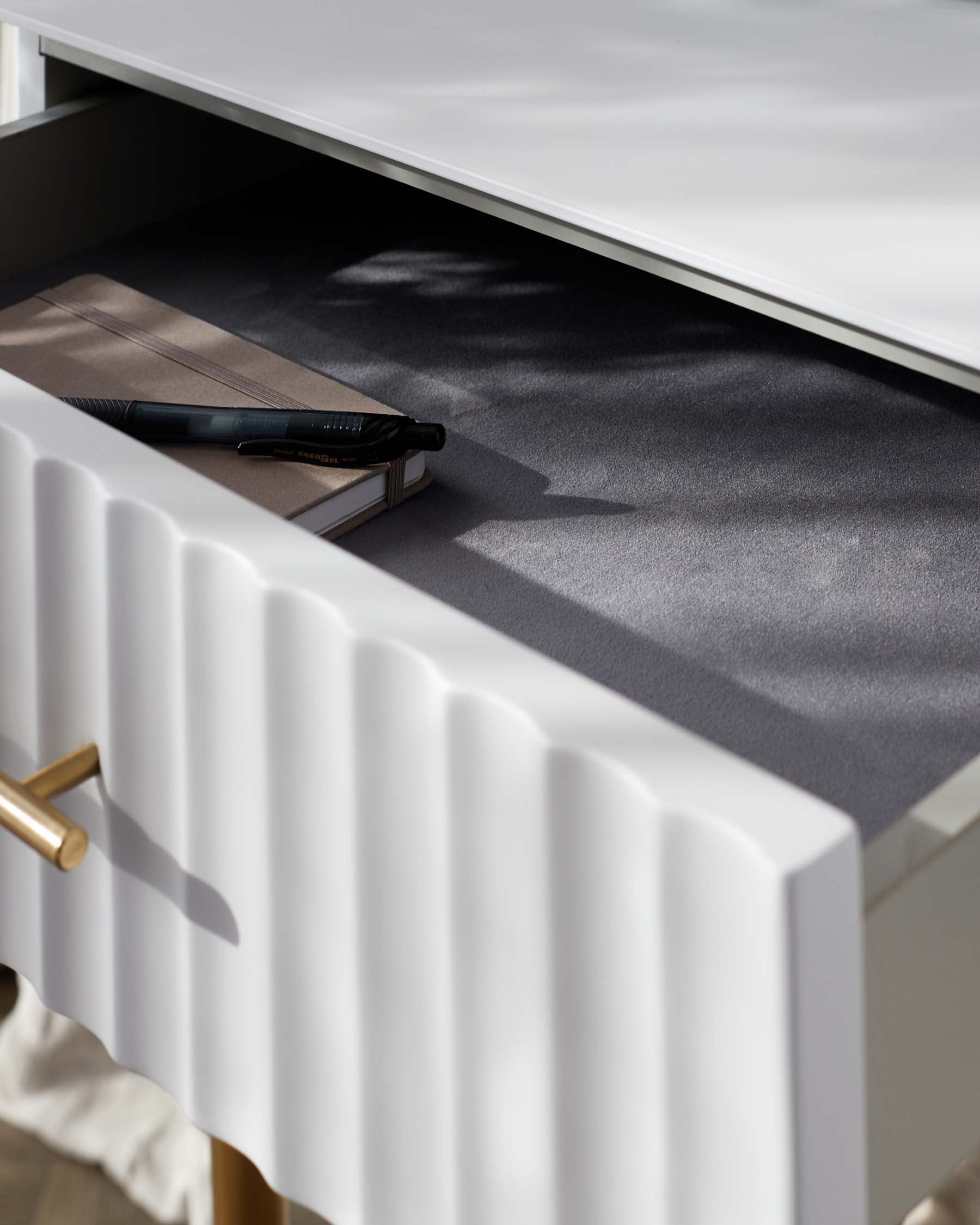 Modern white office desk with sculptural legs and gold drawer handles, showcasing an open drawer with a notebook and pen inside.