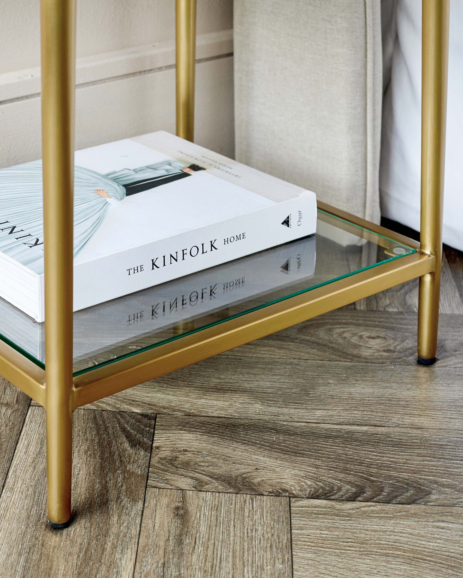 Elegant modern side table with a slender gold-finished metal frame and two glass shelves, featuring a transparent top shelf with books and a reflective mirror bottom shelf. Perfect for adding a touch of luxury and practicality to any living space.