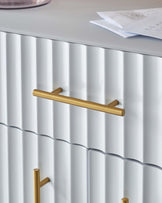 Stylish white cabinet with ribbed texture and gold handles, complemented by a decorative glass vase and notes on top.