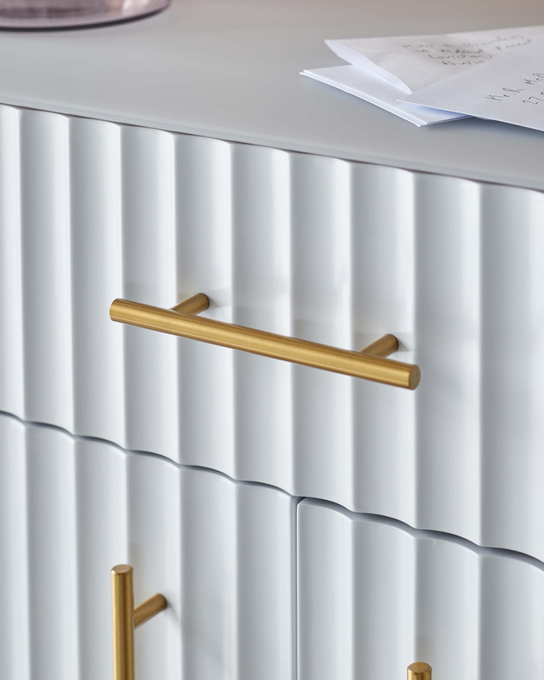 Stylish white cabinet with ribbed texture and gold handles, complemented by a decorative glass vase and notes on top.