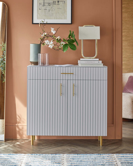 Contemporary gray cabinet with vertical grooves, gold handles, a modern lamp, and decorative vases on a warm-toned wall.